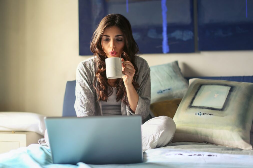 chica sentada sobre una cama con un portátil bebiendo de una taza