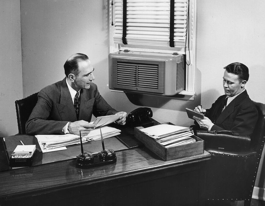Un hombre de negocios toma notas mientras su jefe se sienta detrás de un escritorio y lee las cartas. Un aire acondicionado está montado en la ventana. Foto: Lambert/Getty Images