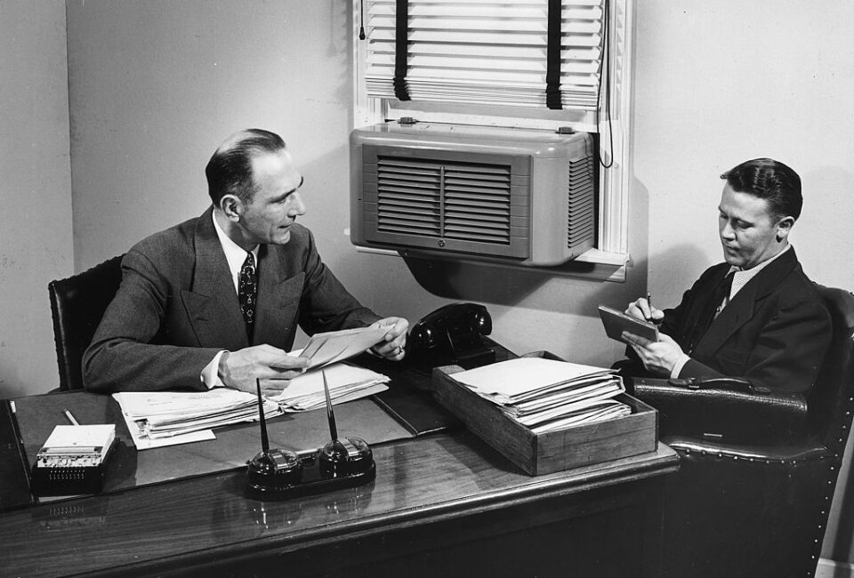 Un hombre de negocios toma notas mientras su jefe se sienta detrás de un escritorio y lee las cartas. Un aire acondicionado está montado en la ventana. Foto: Lambert/Getty Images