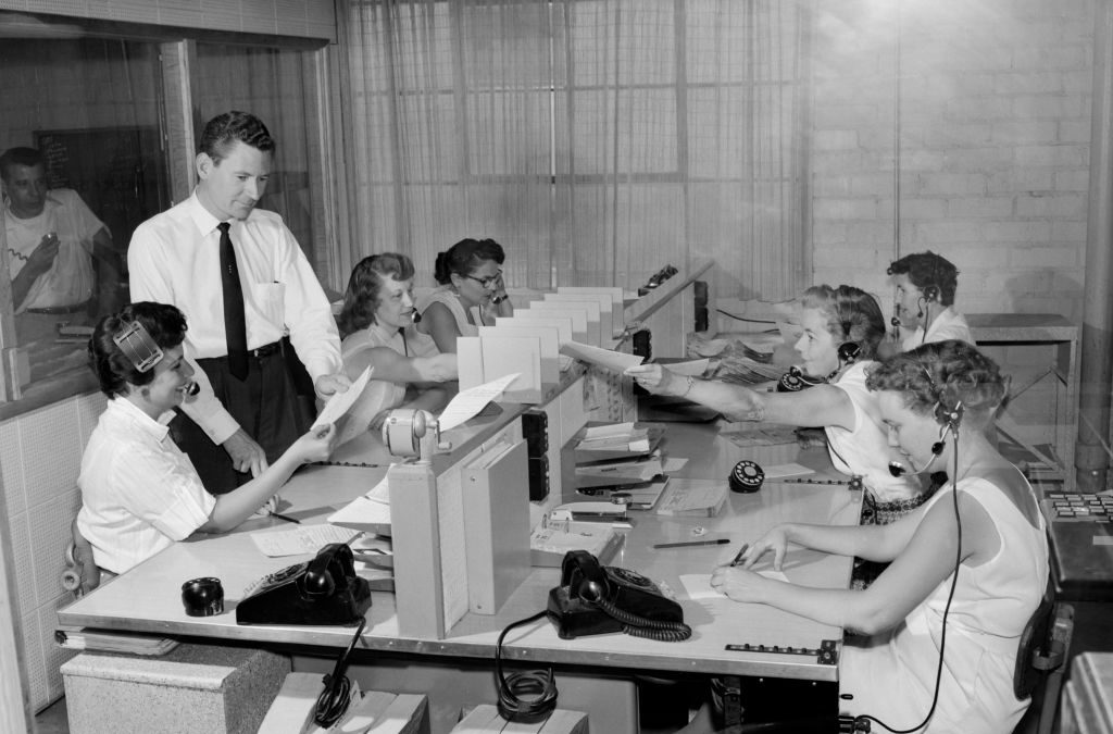 Un grupo de mujeres con auriculares están sentadas en un call center, mientras que un hombre que está de pie revisa unos documentos con una de ellas. Foto: Kirn Vintage Stock/Getty Images